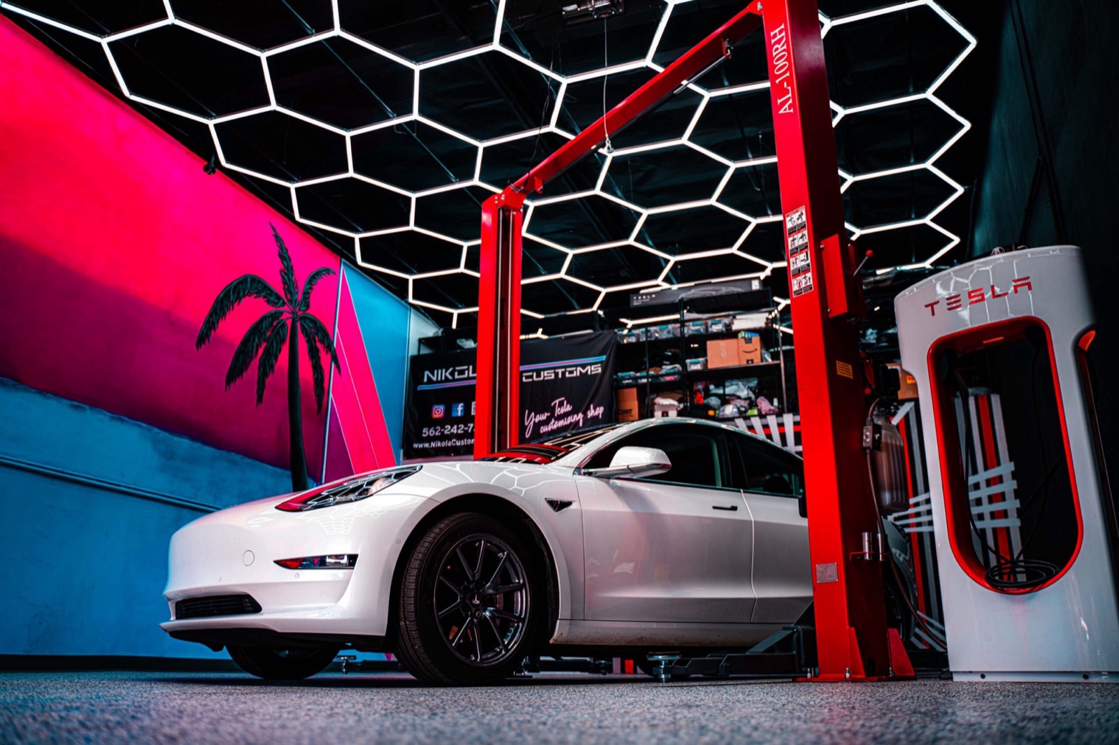 Tesla Model 3 in the Nikola Customs showroom under hex lighting next to a Supercharger